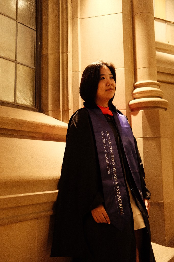 Amy Seunghyun Lee at UW HCDE graduation in a white dress and purple stole, holding a bouquet and mortarboard in front of the Drumheller fountain and brick campus building